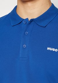 Polo shirt bleu en tissu texturé, avec un col, trois boutons et un logo blanc sur la poitrine.