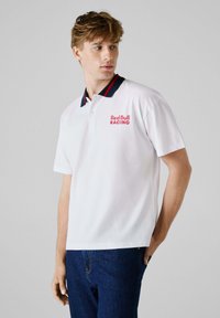 Hvid polo-shirt med korte ærmer, navy- og rødstribet krave, og "Red Bull RACING" logo i rødt på venstre bryst. Blød stoftekstur.