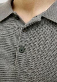 Šedé pletené polo tričko s texturovaným vzorem, které má límec a dva zelené knoflíky u krku. Detailní pohled na látku.