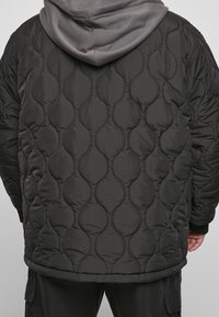 Personne portant une veste matelassée noire avec un sweat à capuche gris en dessous, vue de dos.