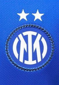 Μπλε αθλητικό ύφασμα με ένα κυκλικό μαύρο και μπλε κεντητό σήμα που φέρει λευκά στιλιζαρισμένα γράμματα και δύο λευκά αστέρια από πάνω.