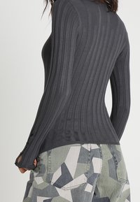 Haut gris à manches longues côtelé en matière extensible, avec des trous pour les pouces et une coupe ajustée, associé à un pantalon à motif géométrique.