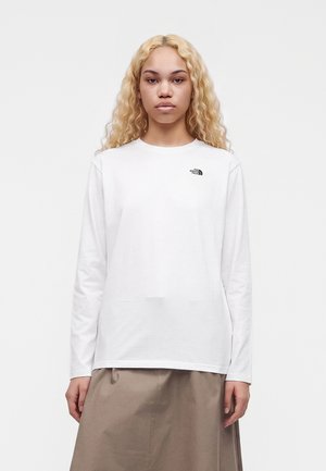 Jeune femme aux longs cheveux blonds bouclés portant un t-shirt blanc à manches longues avec le logo The North Face et une jupe beige, debout devant un fond uni.
