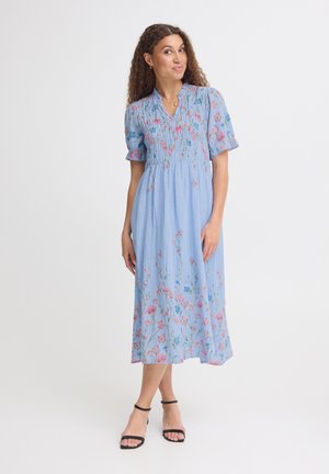 Kvinde iført en lyseblå blomster midi-kjole med puffede ærmer og sorte hælede sandaler står mod en ensfarvet hvid baggrund.