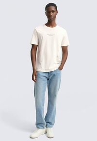 Lysebeige t-shirt med korte ærmer og et lille tryk på brystet, kombineret med lyseblå jeans med lige ben og hvide sneakers.