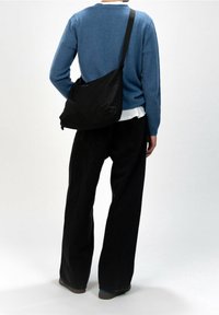 Černá kabelka přes tělo z měkké látky, s měkkým designem, nastavitelným popruhem a minimalistickým logem vpředu.