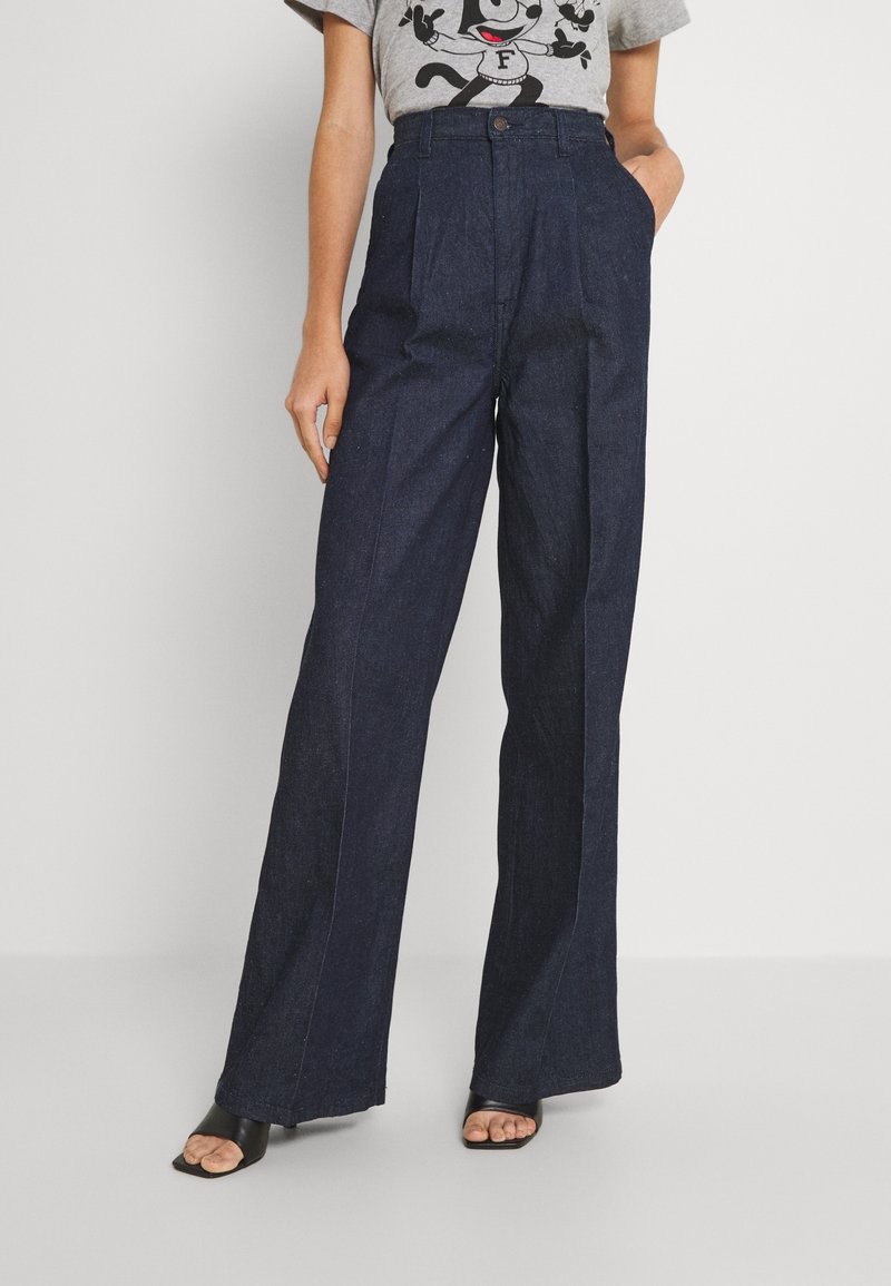 Femme portant un jean large taille haute bleu foncé, un t-shirt gris à motif et des sandales à talons ouvertes noires, debout avec une main dans la poche.