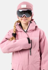 Giacca da sci impermeabile rosa chiaro con colletto alto, dettagli neri e logo. Abbinata a un berretto in maglia rosa coordinato e occhiali da sci.