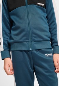 Hummel sporttracksuit in teal en zwart. Bevat een jack met ritssluiting, ribgebreide manchetten en contrasterende witte zijpanelen met logo-informatie.