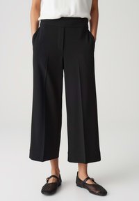 Black wide-leg pants with a seamless waistband, front pleats, side pockets, made from a smooth fabric. Paired with black mesh flats.