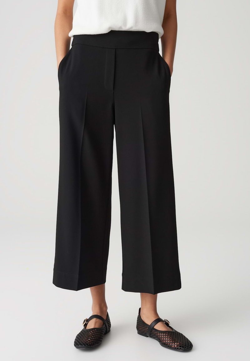 Black wide-leg pants with a seamless waistband, front pleats, side pockets, made from a smooth fabric. Paired with black mesh flats.