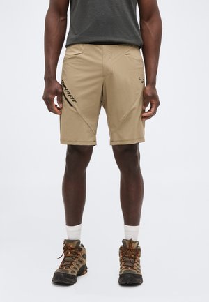 Mężczyzna w beżowych spodenkach outdoor z czarnym logo „DYNAFIT”, białych skarpetkach i butach trekkingowych na jednolitym tle.