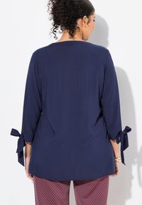Tunic blu navy con maniche a tre quarti, caratterizzata da dettagli con fiocco sui polsini. Texture liscia, design semi-fittato, abbinata a pantaloni con motivo.