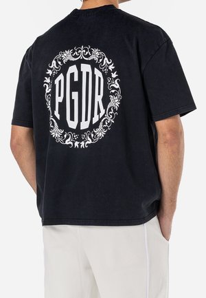 Man wearing a black T-shirt with large white "PGDR" text and decorative floral circle on the back, paired with light-colored pants.