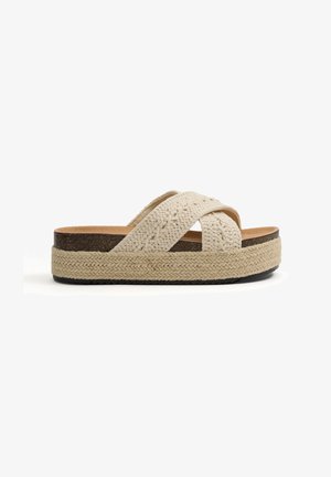 Sandalia tipo slide beige con tiras cruzadas tejidas, plantilla de corcho, entresuela de yute trenzado y suela exterior de goma negra.