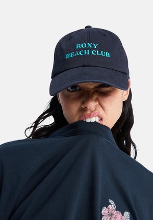 Kobieta w ciemnoniebieskiej czapce z napisem „Roxy Beach Club”, gryząca ciemnoniebieską koszulkę z różowym kwiatowym wzorem, mrugająca ze skrzywioną twarzą.