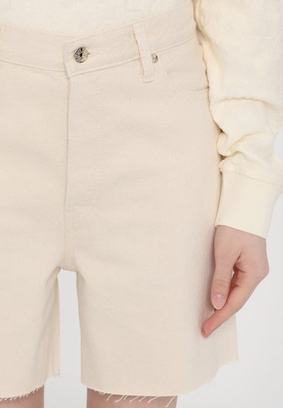 Beige farkkushortsit, joissa on repaleinen helma, yksi napin kiinnitys ja näkyvät etutaskut. Materiaali vaikuttaa pehmeältä ja sileältä.