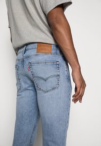 Hombre que lleva pantalones vaqueros Levi's de color azul claro y una camiseta gris, visto desde la cintura hasta la mitad del muslo contra un fondo simple.