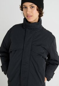 Schwarze Daunenjacke mit hohem Kragen, ausgestattet mit einem Reißverschluss vorne und zwei großen Brusttaschen. Das Material hat eine leicht zerknitterte Textur.