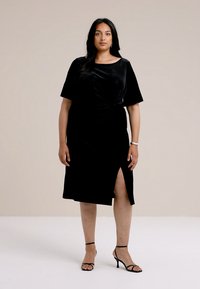 Rochie din catifea neagră cu talie ajustată, mâneci scurte și o fante laterală, având o textură netedă și un design până la genunchi.
