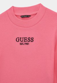 Maglione rosa a coste con scollo rotondo e logo ricamato in nero "GUESS EST.1981" sulla parte anteriore. Tessuto morbido, design casual.