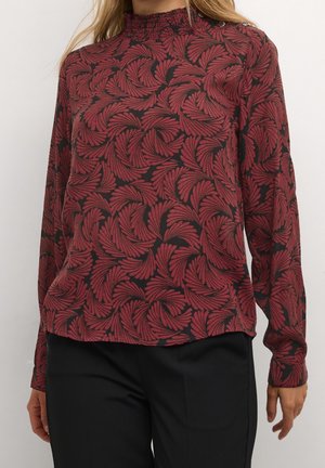 Kvinde iført en bordeaux og sort langærmet top med snurrende bladmønstre og sorte bukser, stående mod en ensfarvet baggrund.