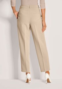 Vrouw in beige wijde broek met witte hoge hakken, staande met één hand in de zak tegen een effen achtergrond.