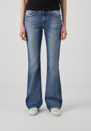 Personne portant un jean bootcut bleu et des baskets blanches, debout, les bras détendus le long du corps, sur un fond clair uni.