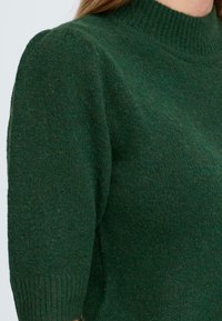 Pull en tricot vert avec un col côtelé haut et des manches courtes. Le matériau a une finition douce et texturée avec de légères variations de couleur.