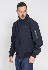 Marineblaue Bomberjacke mit hohem Kragen, Reißverschluss, gerippten Bündchen und einer Reißverschlusstasche am Ärmel. Mit einem dezenten Logo auf der Brust.