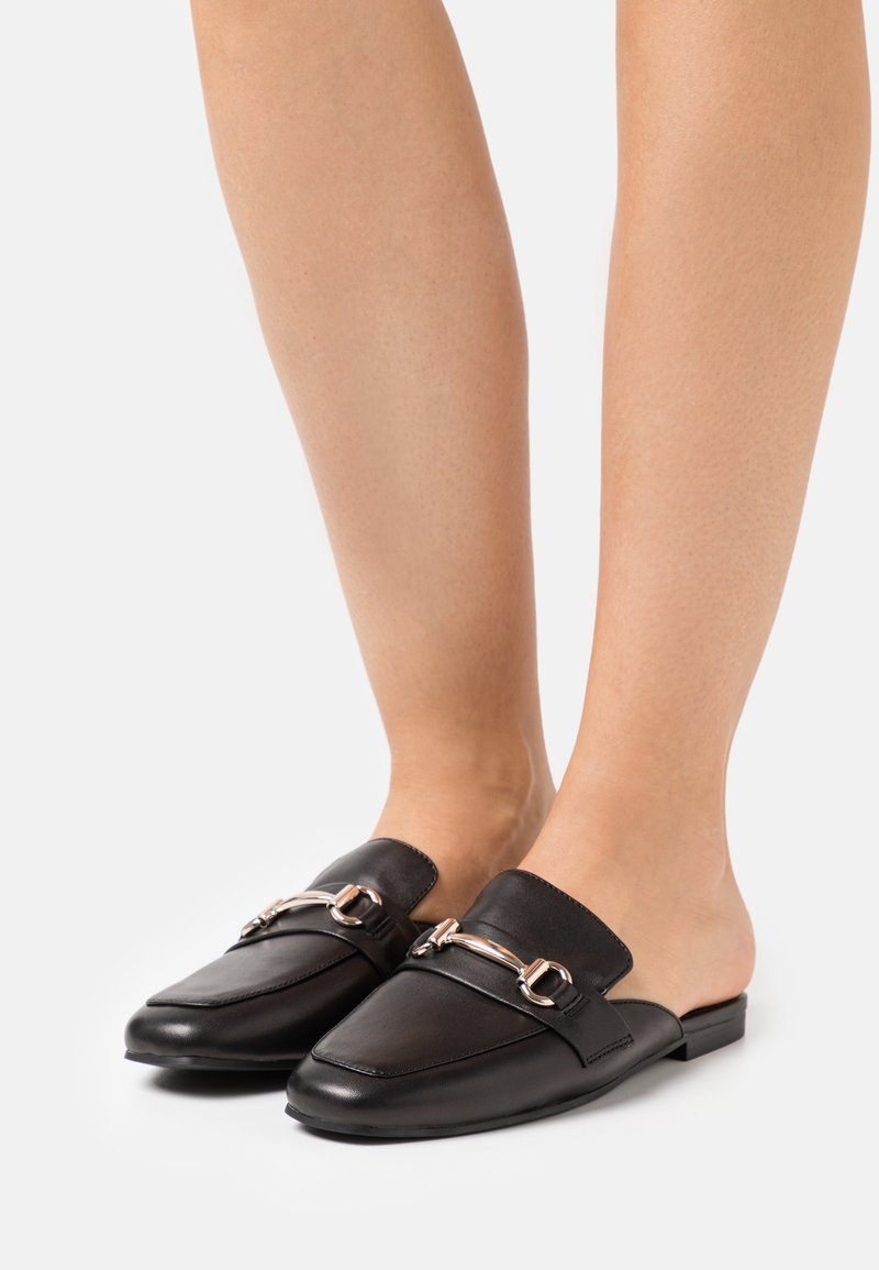 Mocassins noirs en cuir à enfiler avec détail en métal portés pieds nus sur un fond blanc uni.