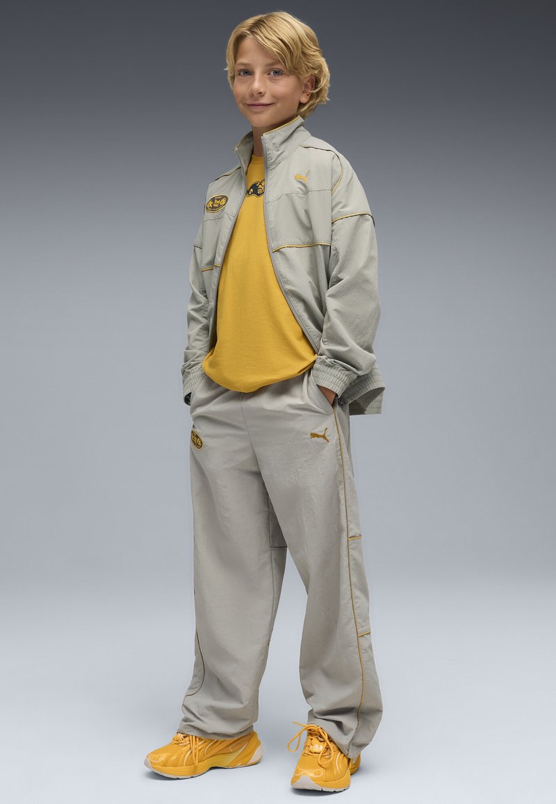 Jasnoszary dres z żółtymi akcentami, składający się z zapinanej na zamek kurtki i luźnych spodni, w połączeniu z jasnosłonecznymi sneakersami.