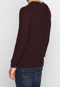 Bordowy sweter typu pulower o teksturowanej powierzchni, z długimi rękawami i zaokrąglonym dekoltem, zestawiony z niebieskimi dżinsami z tylnymi kieszeniami.