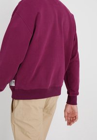 Fialová mikina s volnějším střihem, žebrovanými manžetami a spodním lemem. Hladká textura. Na spodní levé straně jsou viditelné cedulky.