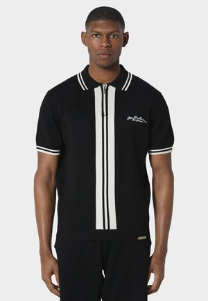 Man draagt een zwart polo met korte mouwen met witte verticale strepen in het midden en witte bies aan de kraag en mouwen.