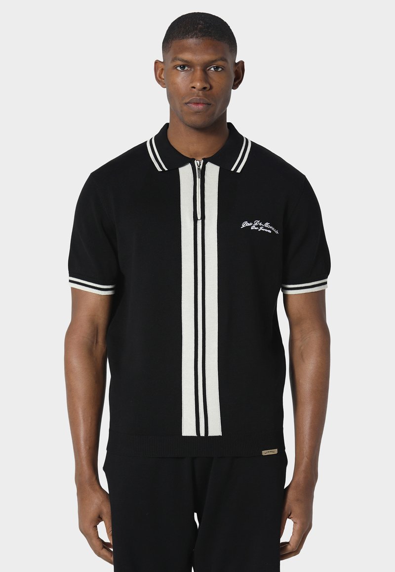 Homme portant un polo noir à manches courtes avec des rayures verticales blanches au centre et des bordures blanches au col et aux manches.