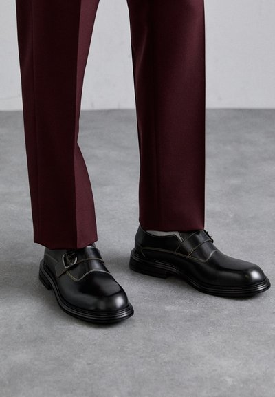 Chaussures en cuir noir avec un détail de boucle, présentant une finition lisse et des coutures contrastantes. Associées à un pantalon bordeaux sur un fond gris.