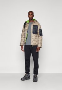 Puffer jacket in beige with textured pattern, green accents, and navy patch pockets. Paired with a gray shirt and black pants. Running shoes featured.