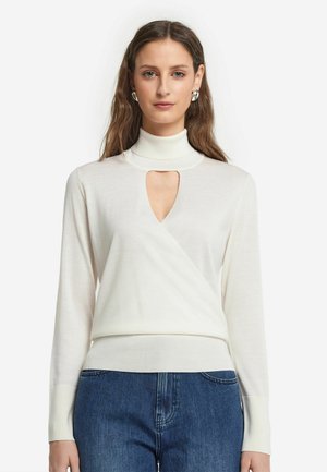 Pull léger blanc avec un col montant, découpe en forme de clé, design croisé et ourlet côtelé. Modèle associé à un jean bleu.