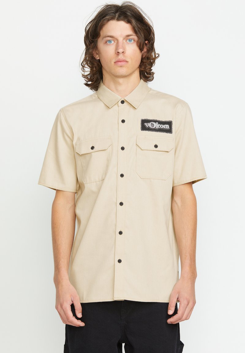 Beige kortærmet skjorte med knapper og to forlommer, sorte knapper og et sort logo-patch på venstre bryst. Flad krave-design.