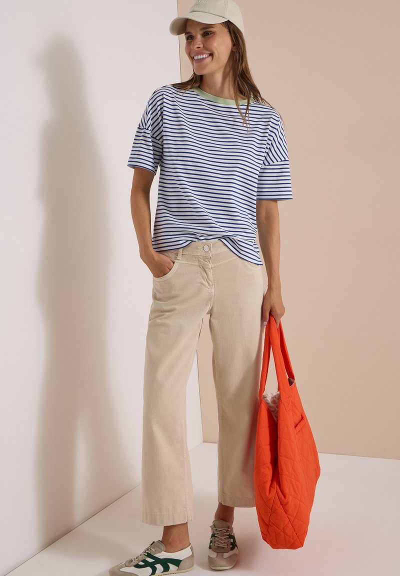 Femme portant une casquette beige, une chemise rayée bleu et blanc, un pantalon beige, des baskets, tenant un sac fourre-tout matelassé orange.