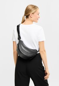 Borsa a tracolla in pelle nera con forma curva, chiusura con zip e tracolla regolabile, indossata sopra una maglietta bianca, con una texture liscia.