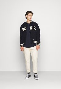 Giacca varsity navy con scritta "DC KIES", polsini a righe, t-shirt bianca, pantaloni color crema chiaro e sneakers nere alte in un ambiente neutro.