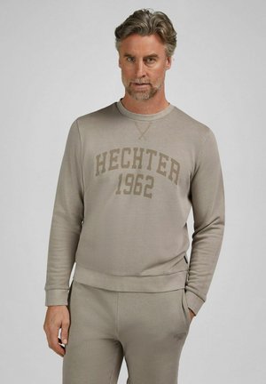 Mann mit grauem Haar und Bart, trägt einen beigen Pullover und passende Hose mit dem Aufdruck "HECHTER 1962" auf dem Oberteil.