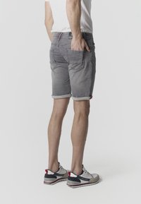 Mand iført op-rullede grå denimshorts og hvide sneakers, stående med den ene hånd i baglommen mod en grå baggrund.