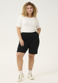 Shorts neri in cotone con vita elasticizzata e tasche laterali, abbinati a una maglietta bianca a maniche corte a costine. Il modello indossa sneaker bianche.