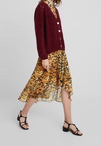 Burgundy knitted cardigan with a V-neck and three buttons, paired with a flowing yellow and brown patterned dress and black strappy sandals.