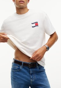 Άνδρας που φοράει λευκό T-shirt Tommy Jeans και μπλε τζιν σηκώνει το πουκάμισο για να δείξει την κοιλιά, φορά ασημένιο ρολόι χειρός και μαύρη ζώνη.