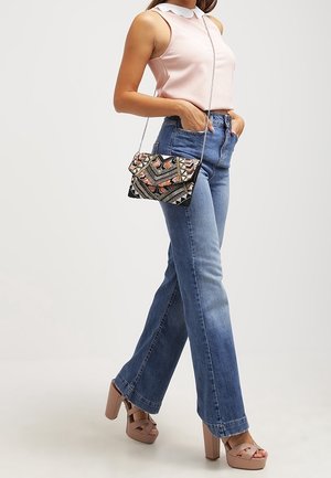 Donna che indossa un top senza maniche rosa, jeans blu a vita alta, tacchi a piattaforma nude e porta una borsa a tracolla con motivo.