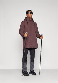 Bordoizolovaná bunda s kapucňou, dve predné vrecká, v kombinácii s čierno-sivými nohavicami. Drží turistické palice, má na nohách čiernu obuv a slnečné okuliare.